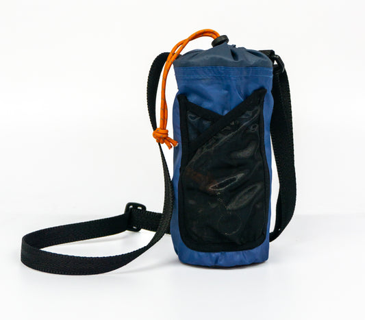 The image is of a navy blue bottle holder made from salvaged tent material. The bottle holder has a black mesh pocket on the front, a black draw cord with an orange puller closure at the top, and a black webbing strap.