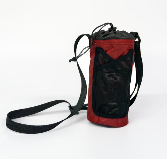 The image is of a red bottle holder made from salvaged tent material. The bottle holder has a black mesh pocket on the front, a black draw cord closure at the top, and a black webbing strap.