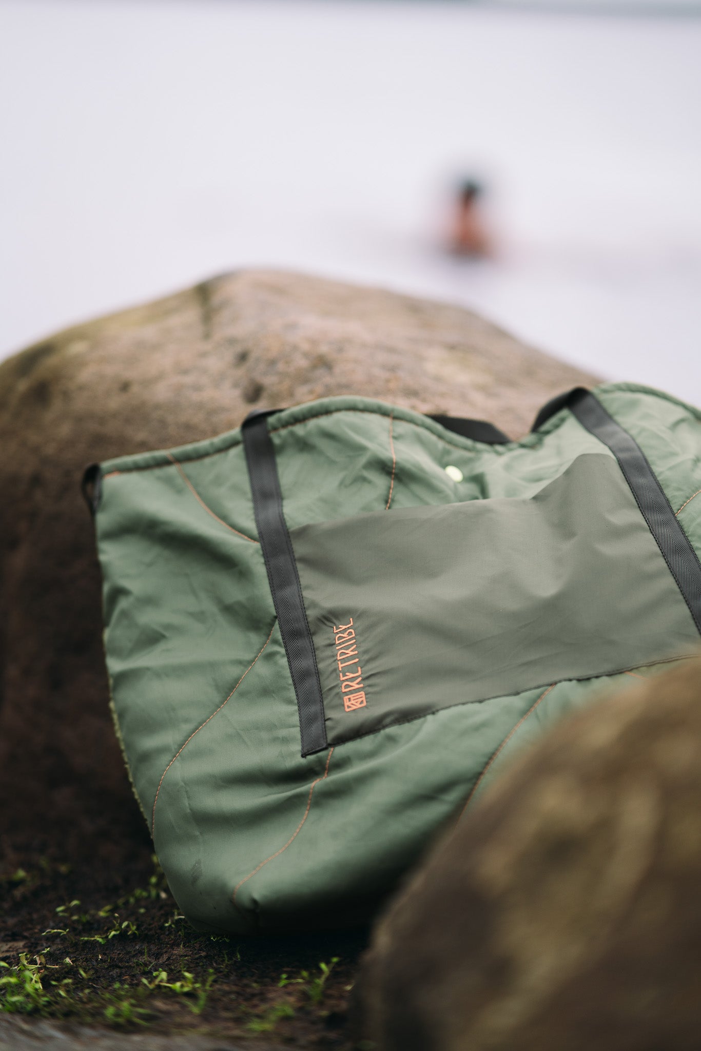 Remade tote bag, made from salvaged sleeping bags. With a front pocket made from salvaged green tent material. Embroidered with the Retribe logo vertically in the left hand side corner. Black webbing straps make the bag stable and strong. The bag closes with a popper in the centre. The bag is photographed on a rock next to a reservoir where a women is open water swimming in the winter weather with a black fluffy hat on.